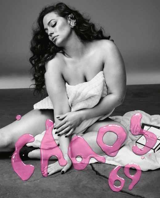 Ashley Graham poses wrapped in a towel next to pink, fluid typography and a Chanel box on a grey surface in an indoor studio setting.