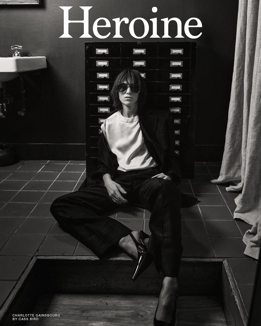 Black-and-white fashion shot of Charlotte Gainsbourg sitting on a tiled floor wearing a jacket, pants, blouse, and sunglasses.