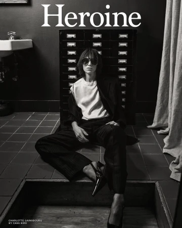 Black-and-white fashion shot of Charlotte Gainsbourg sitting on a tiled floor wearing a jacket, pants, blouse, and sunglasses.
