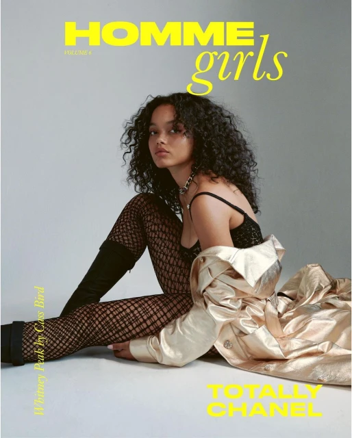 Fashion magazine cover featuring a woman with curly hair, wearing a black top, fishnet tights, and a gold coat, posing against a gray background.
