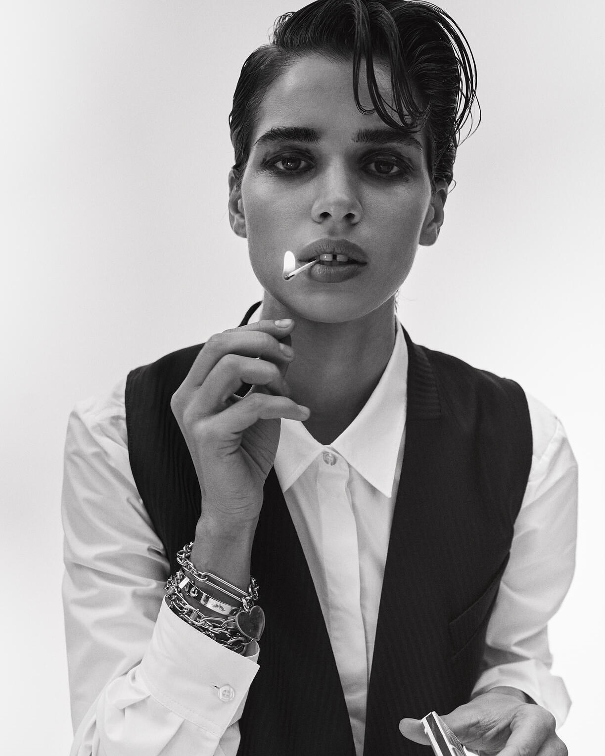 Monochrome portrait of a woman with short dark hair, wearing a white shirt and vest, holding a lit match in her mouth.