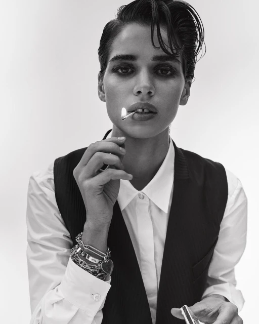 Monochrome portrait of a woman with short dark hair, wearing a white shirt and vest, holding a lit match in her mouth.