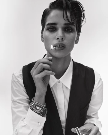 Monochrome portrait of a woman with short dark hair, wearing a white shirt and vest, holding a lit match in her mouth.