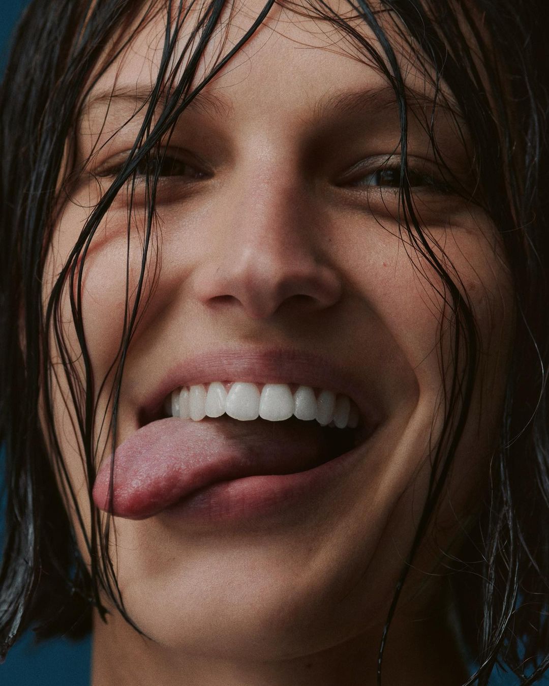 Close-up of a smiling woman with wet dark hair sticking her tongue out, against a blue background, showcasing a youthful and playful expression.