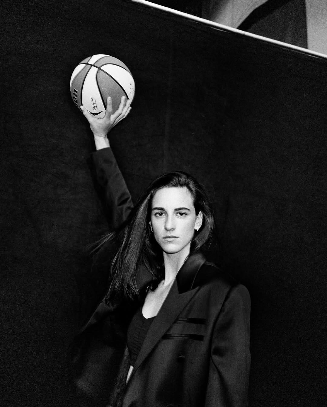 Portrait of a woman wearing a dark blazer holding a basketball overhead against a dark background