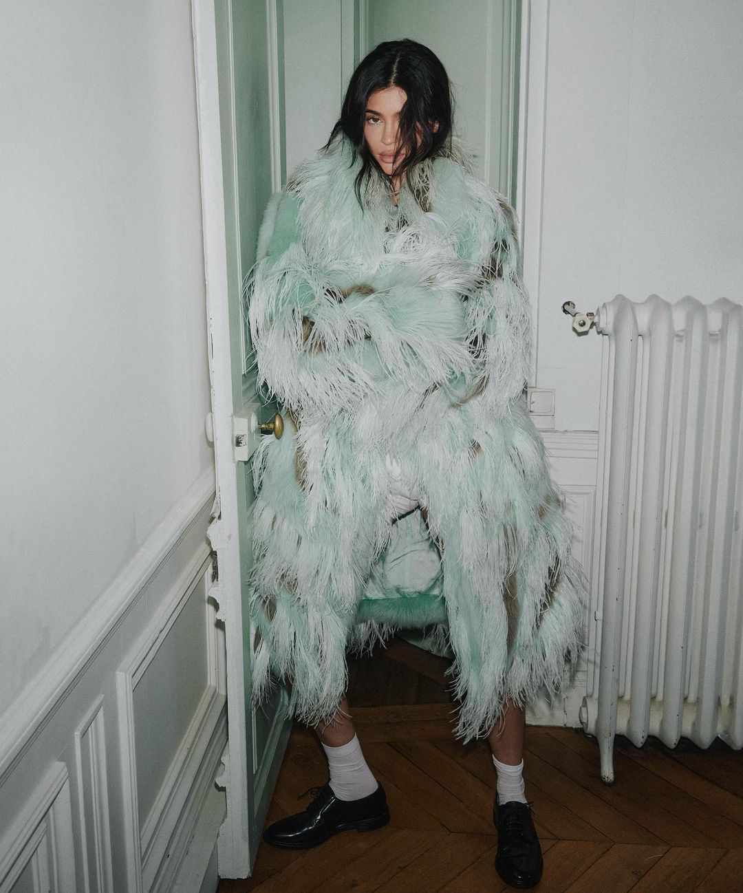 A young adult woman poses in an ornate, feathered coat in a stark white interior setting with white trim, door and radiator.