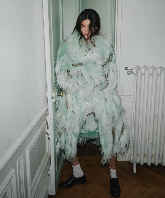 A young adult woman poses in an ornate, feathered coat in a stark white interior setting with white trim, door and radiator.