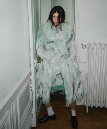 A young adult woman poses in an ornate, feathered coat in a stark white interior setting with white trim, door and radiator.