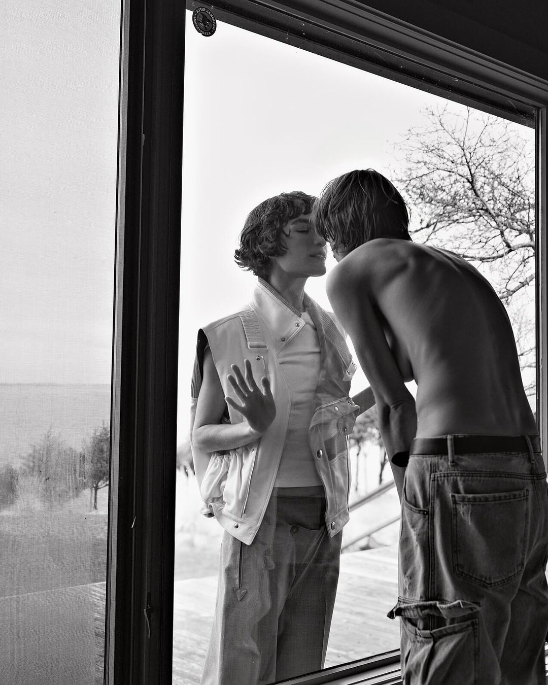 Black and white portrait of a couple looking at each other through a window, showcasing minimalist styling and natural light.