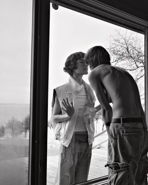 Black and white portrait of a couple looking at each other through a window, showcasing minimalist styling and natural light.