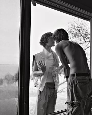 Black and white portrait of a couple looking at each other through a window, showcasing minimalist styling and natural light.
