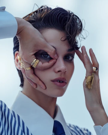 Close-up of a young adult female model with short dark hair and gold rings, posing with her hands around her eye in a studio.