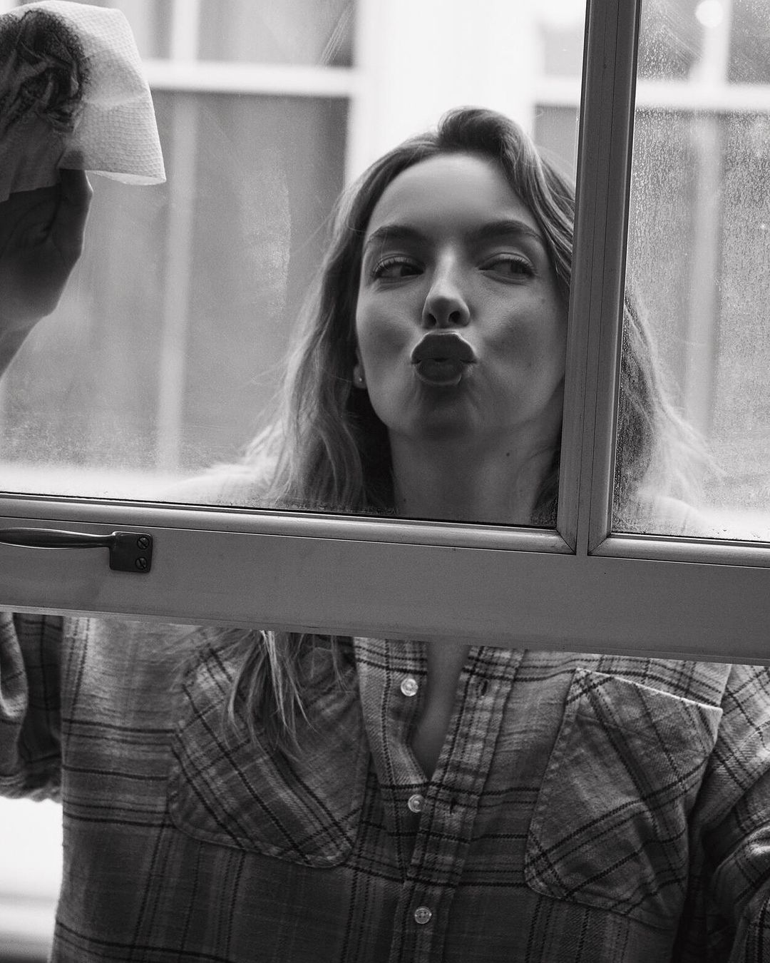 A young adult woman with long hair, wearing a flannel shirt, cleans a window with a paper towel and makes a kissing face in a monochrome portrait.