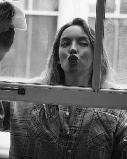 A young adult woman with long hair, wearing a flannel shirt, cleans a window with a paper towel and makes a kissing face in a monochrome portrait.