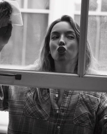 A young adult woman with long hair, wearing a flannel shirt, cleans a window with a paper towel and makes a kissing face in a monochrome portrait.
