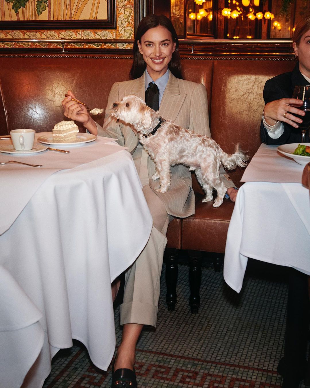Irina Shayk poses indoors at a dining table with a small dog, wearing a pinstripe suit, tie, and loafers while eating cake.