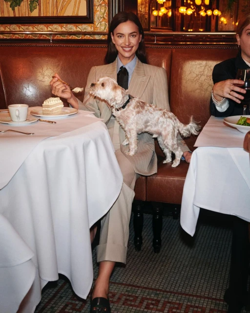 Irina Shayk poses indoors at a dining table with a small dog, wearing a pinstripe suit, tie, and loafers while eating cake.