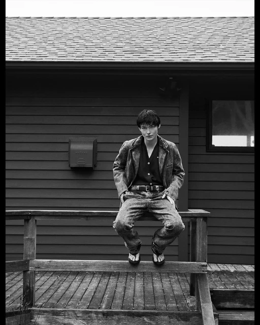 Monochrome portrait of a person wearing a blazer and jeans while sitting on a wooden porch with a house as a backdrop.