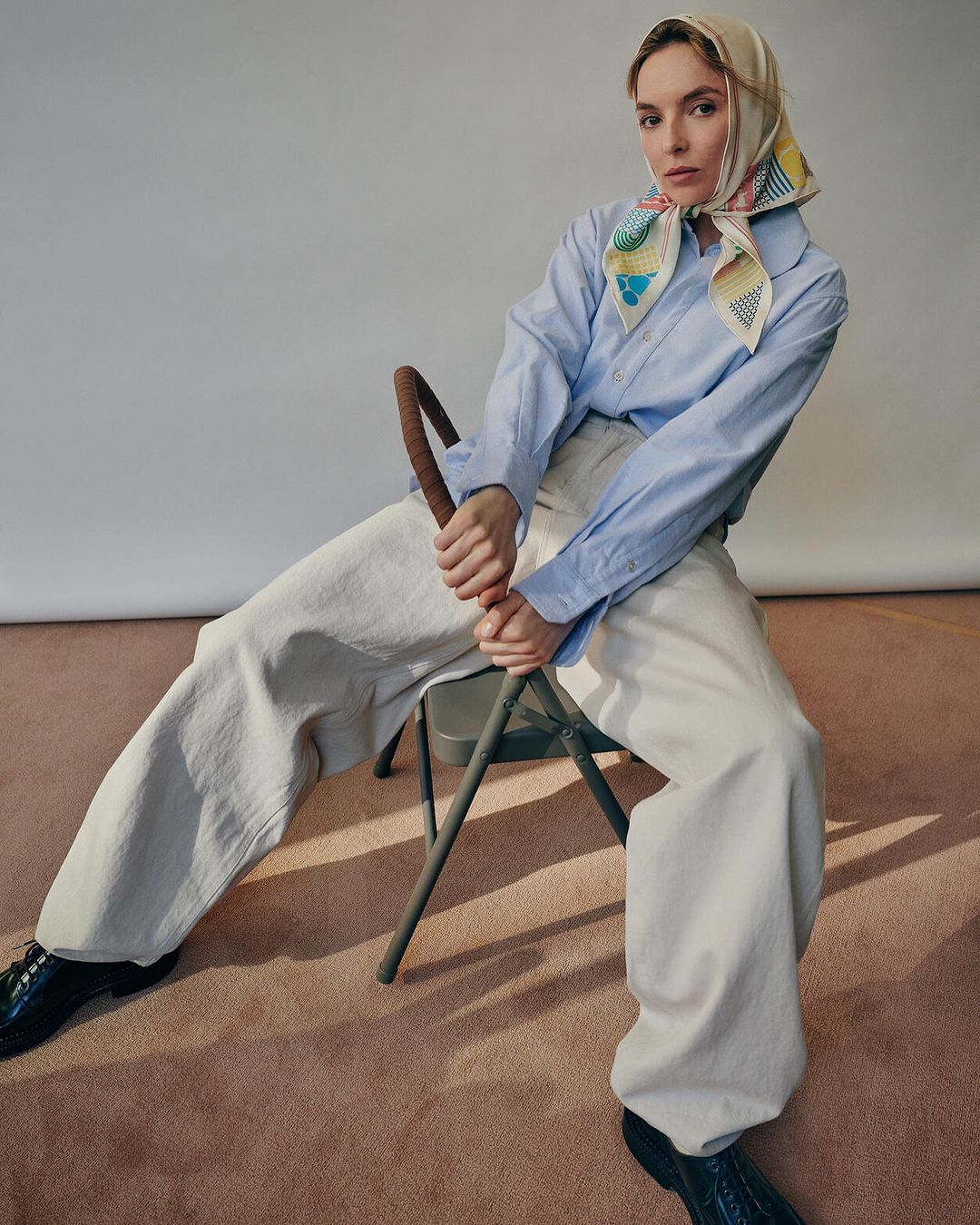Elegant woman sits on a chair, wearing a light blue shirt, cream-colored pants, black patent shoes, and a patterned headscarf in a studio setting.