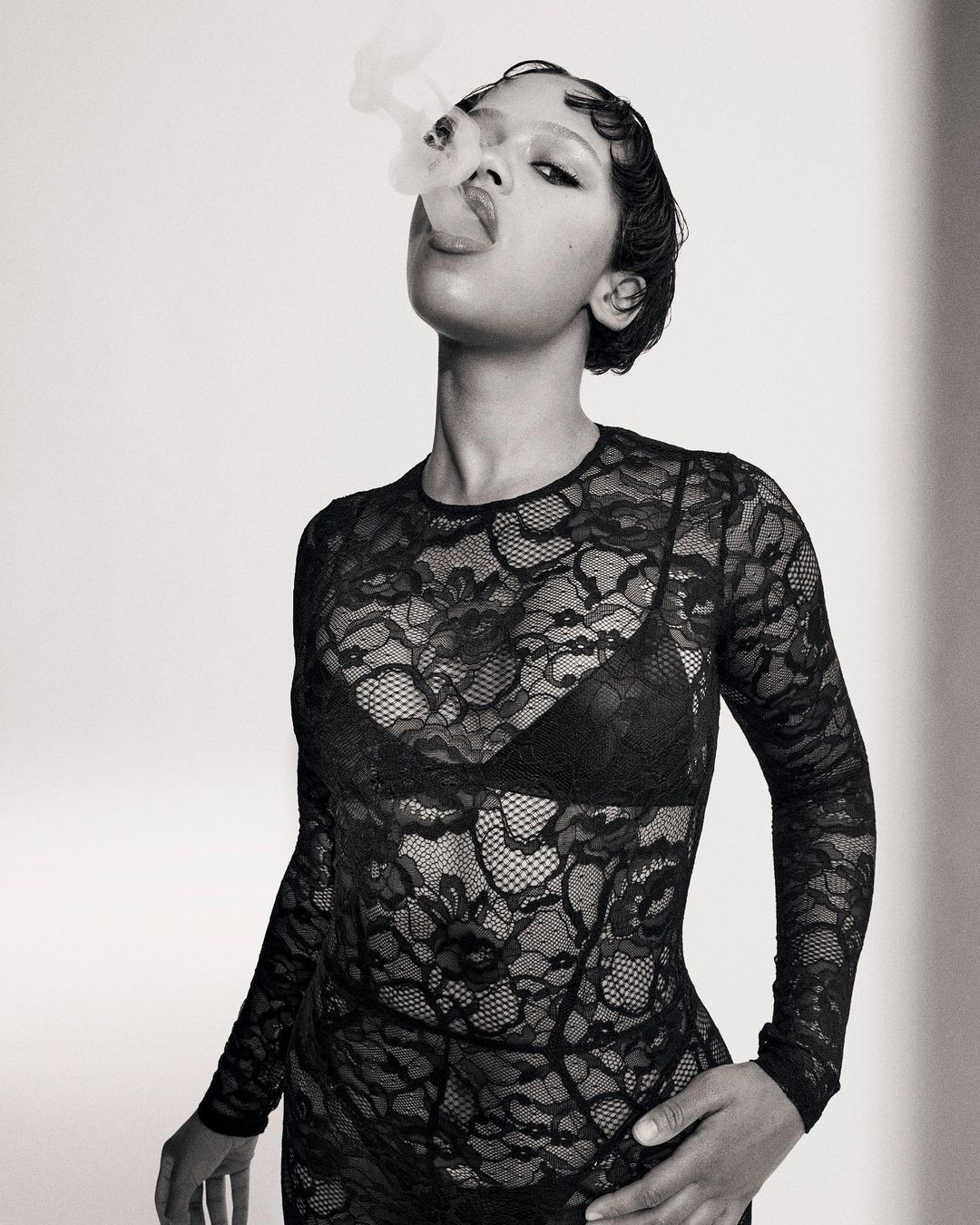 Portrait of a Black female adult exhaling smoke, wearing a black lace top and bra, short dark hair, against a neutral background.