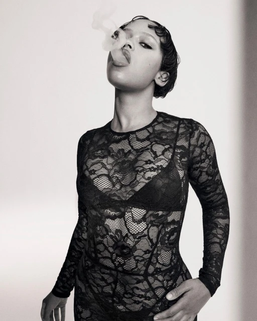 Portrait of a Black female adult exhaling smoke, wearing a black lace top and bra, short dark hair, against a neutral background.