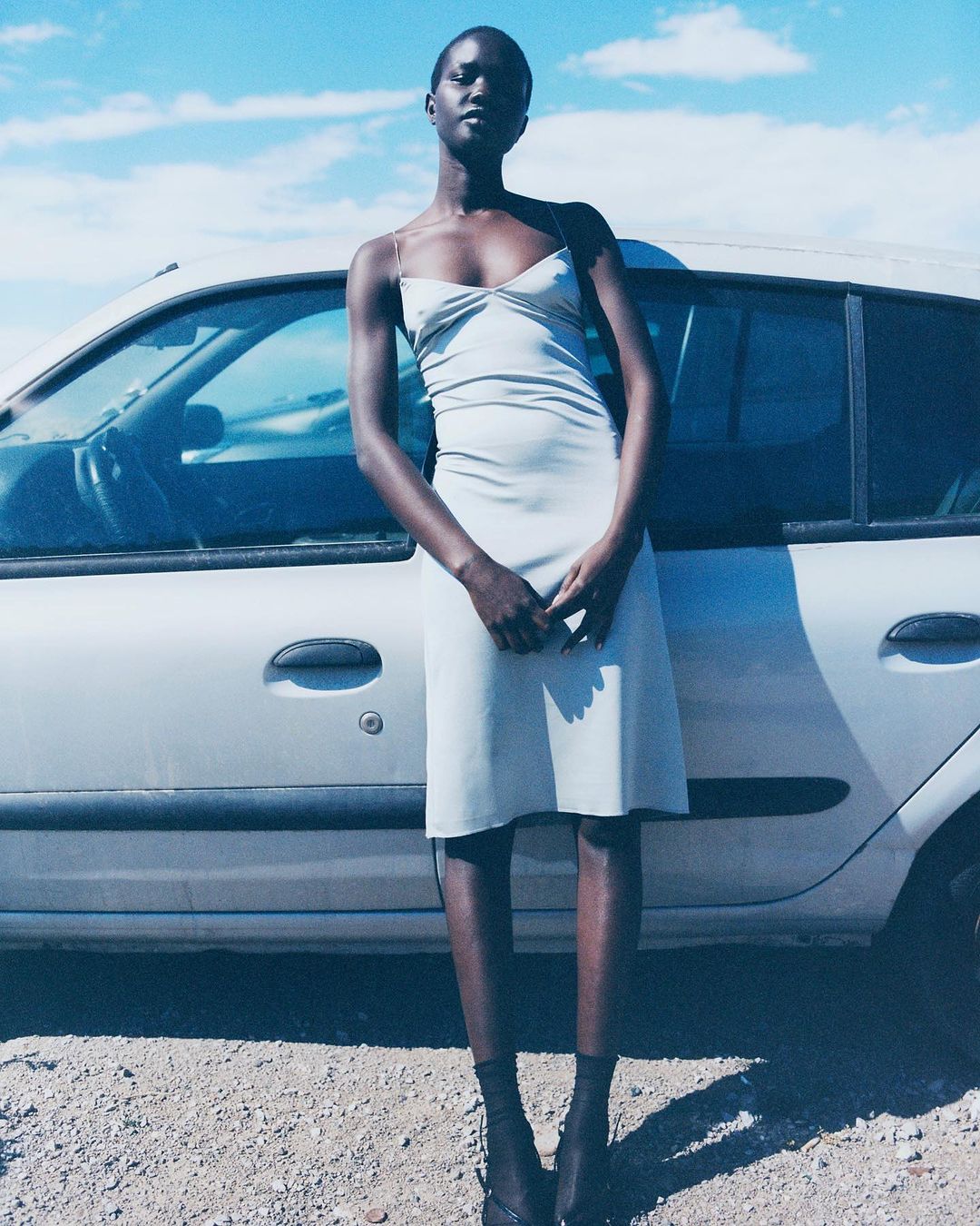 An African woman stands in front of a silver car, wearing a slip dress, in a daytime outdoor setting with a cloudy blue sky.