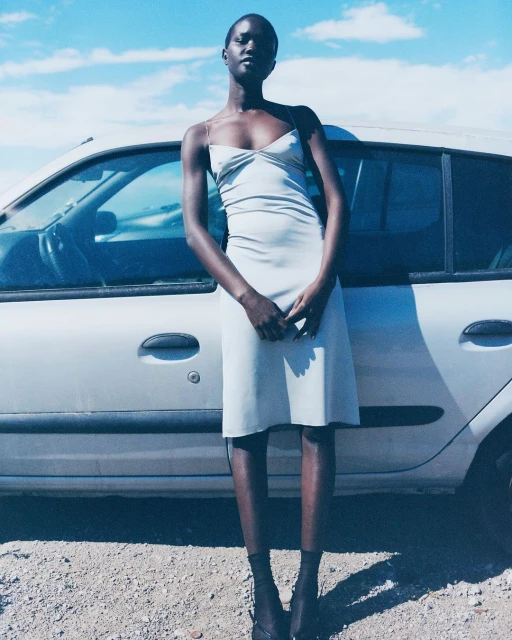 An African woman stands in front of a silver car, wearing a slip dress, in a daytime outdoor setting with a cloudy blue sky.
