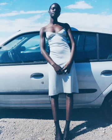 An African woman stands in front of a silver car, wearing a slip dress, in a daytime outdoor setting with a cloudy blue sky.