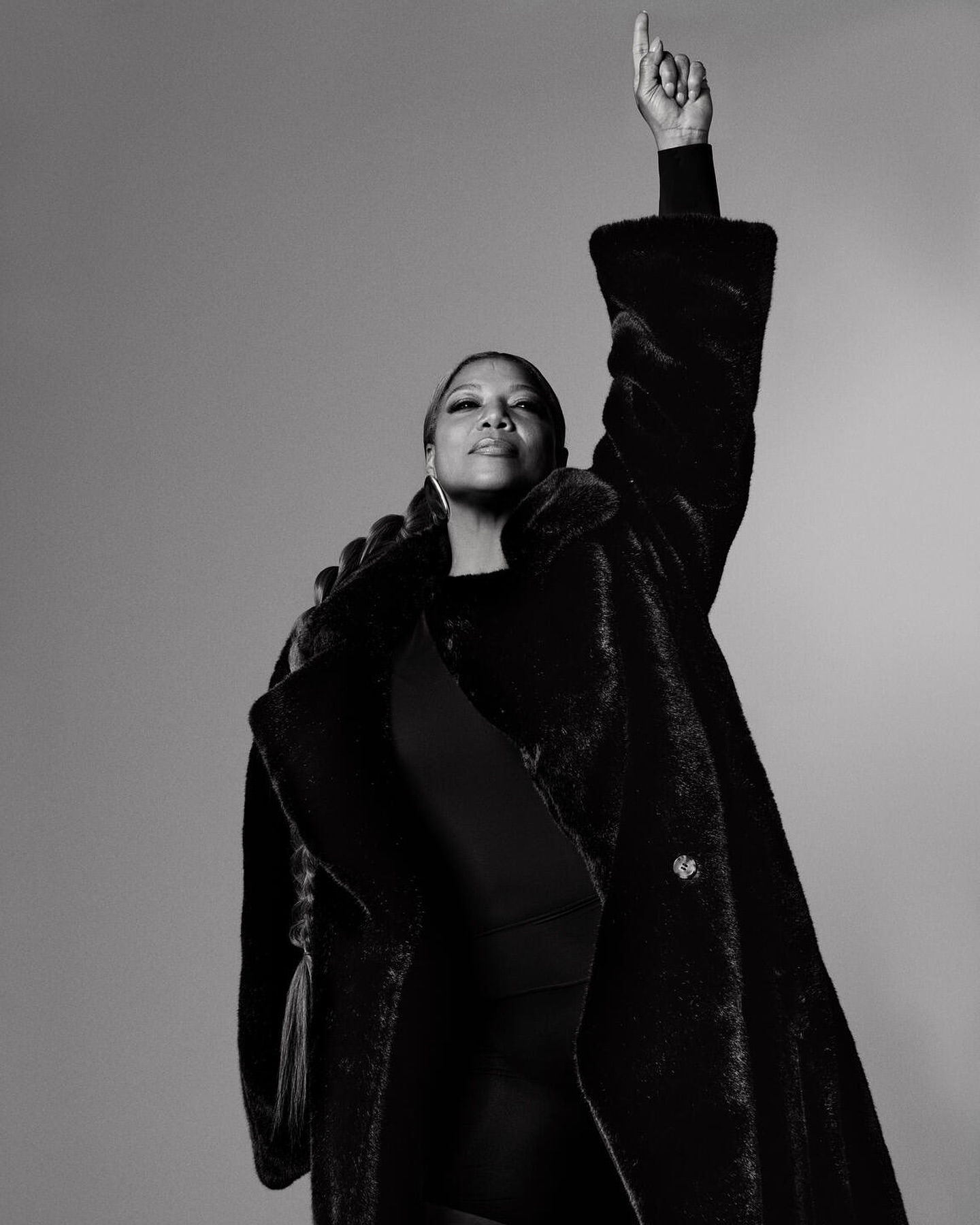 Black and white portrait of an adult woman with a raised hand wearing a fur coat in front of a plain grey background.