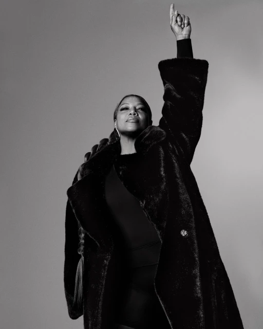 Black and white portrait of an adult woman with a raised hand wearing a fur coat in front of a plain grey background.
