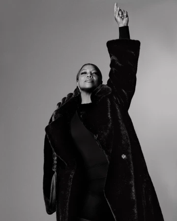 Black and white portrait of an adult woman with a raised hand wearing a fur coat in front of a plain grey background.