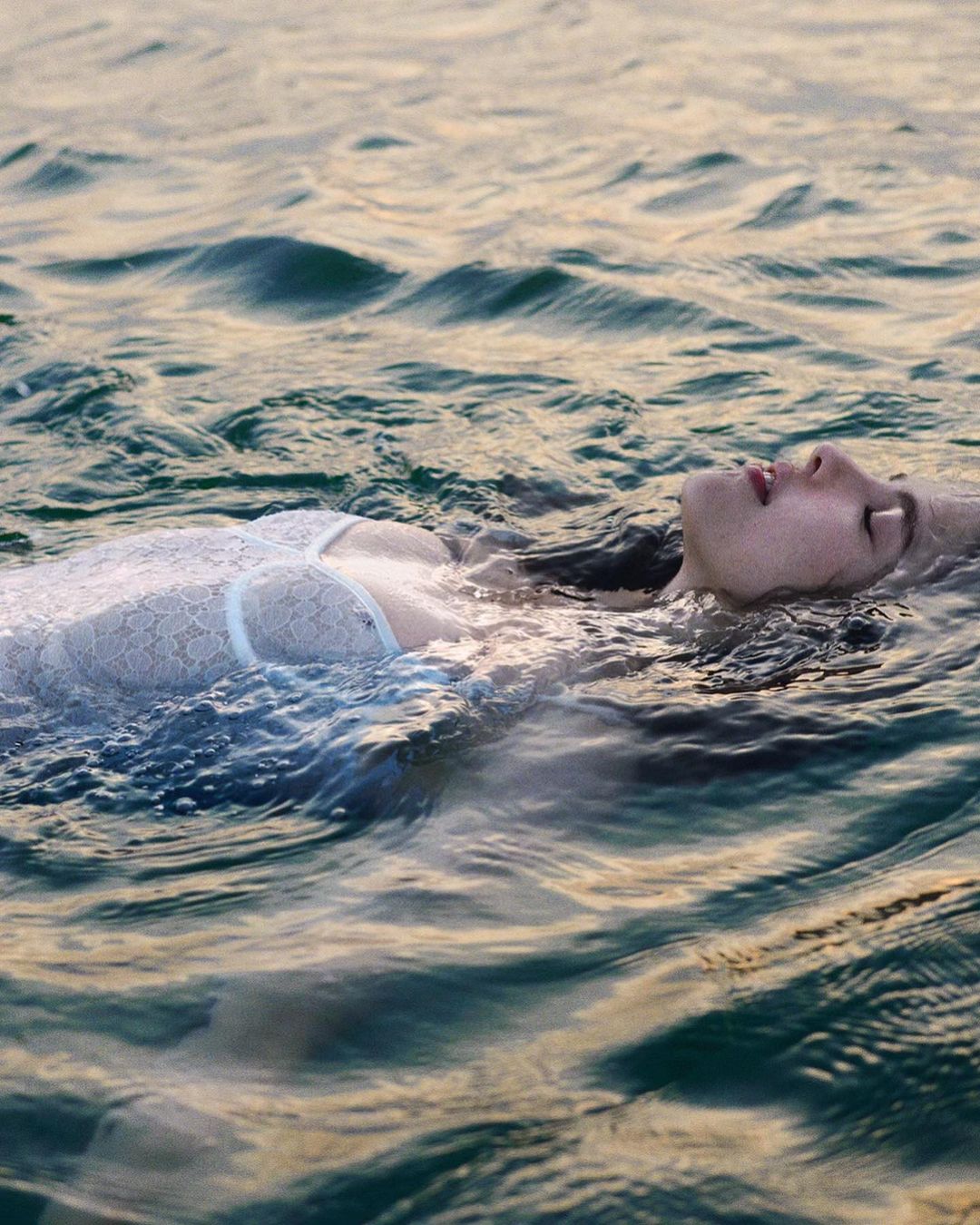 Woman with wet hair floats on her back in ocean water wearing white lace top during the daytime; the water is textured with small waves.