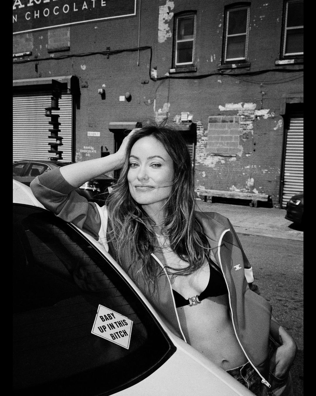 Black and white shot of Olivia Wilde leaning on a car in an urban setting with a building, and a sign that reads 'Chocolate'.