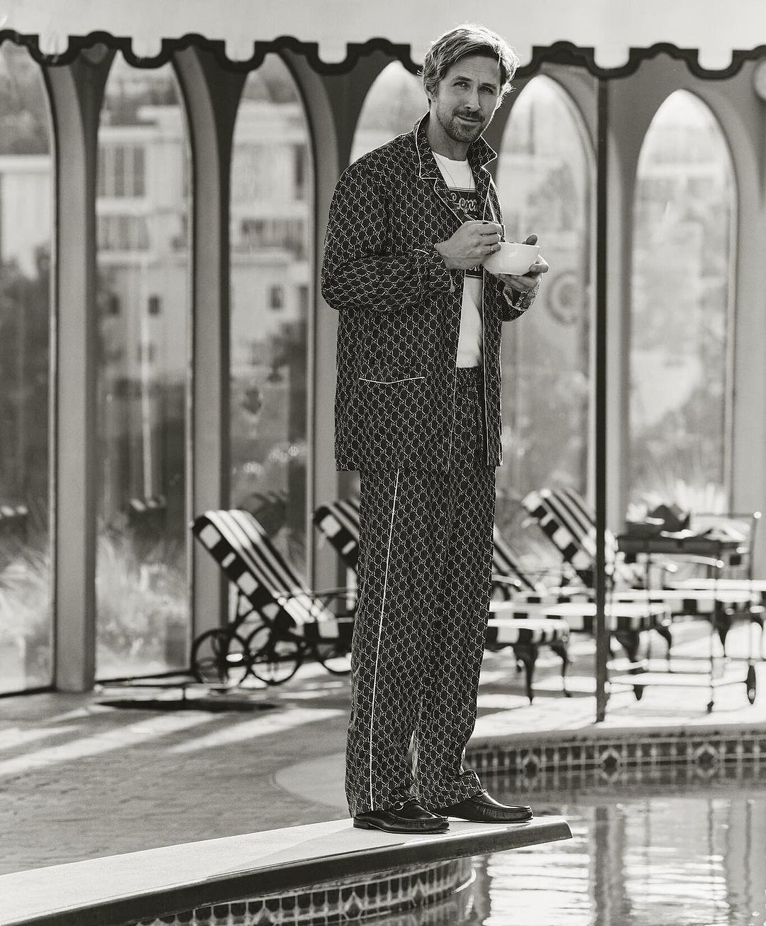 Black and white portrait of a blond male model on a diving board wearing a pajama set holding a teacup near a pool in an arched room.