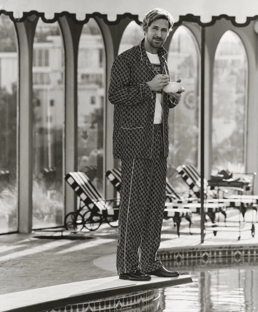 Black and white portrait of a blond male model on a diving board wearing a pajama set holding a teacup near a pool in an arched room.