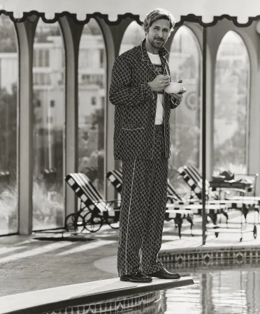 Black and white portrait of a blond male model on a diving board wearing a pajama set holding a teacup near a pool in an arched room.