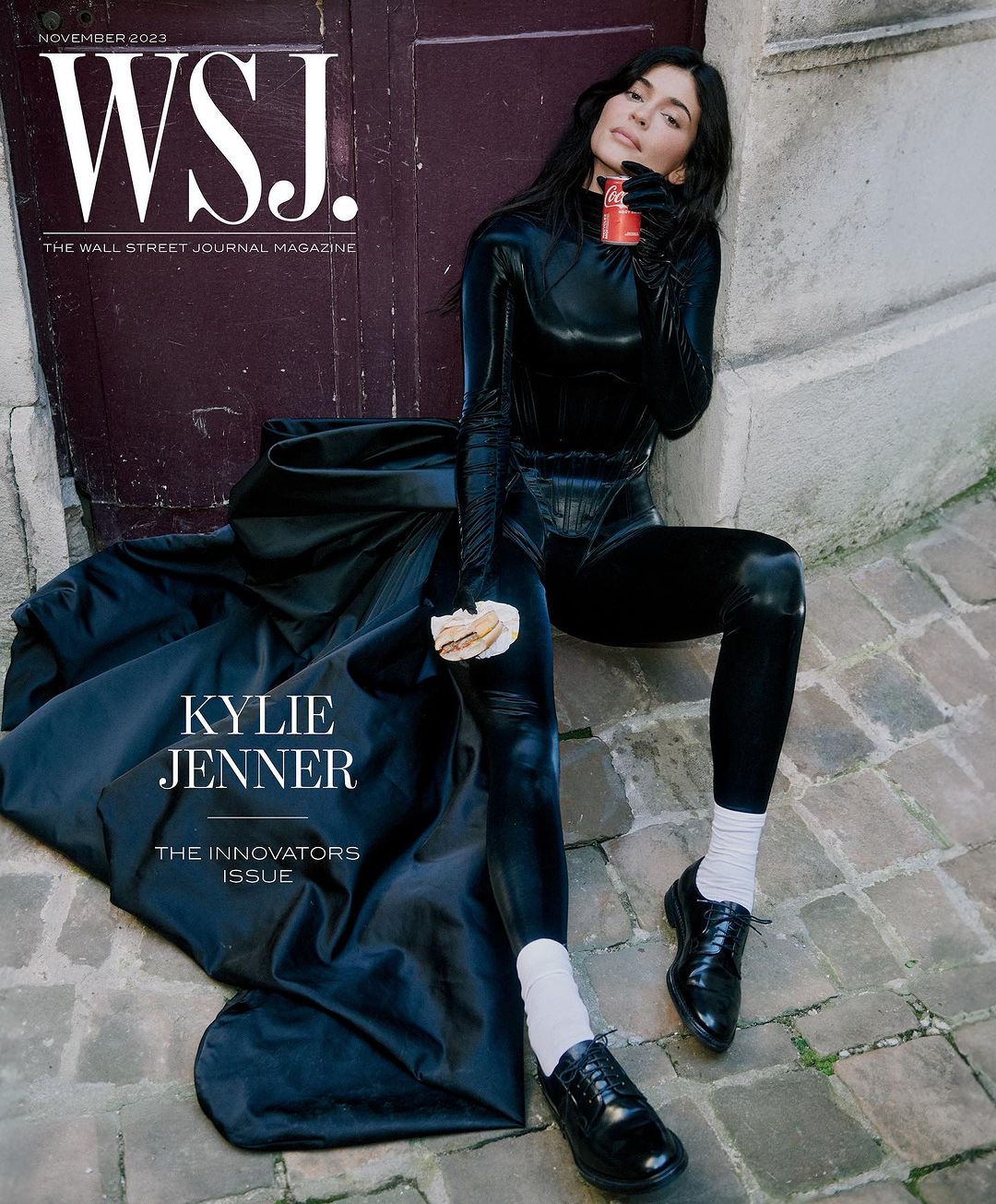WSJ magazine cover featuring Kylie Jenner in a black bodysuit and black coat sitting on stone steps, holding a drink and pastry.