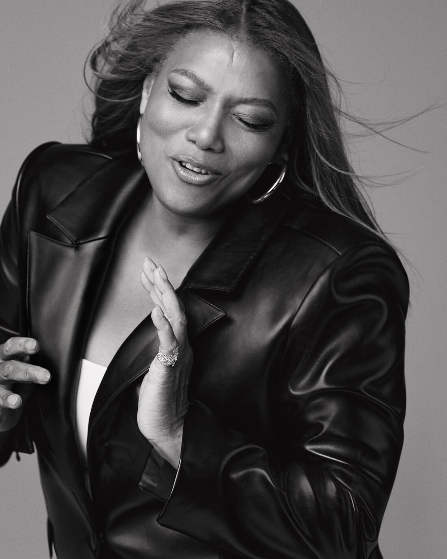 Black and white portrait of Queen Latifah wearing a leather jacket and hoop earrings against a neutral grey background.