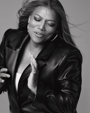 Black and white portrait of Queen Latifah wearing a leather jacket and hoop earrings against a neutral grey background.