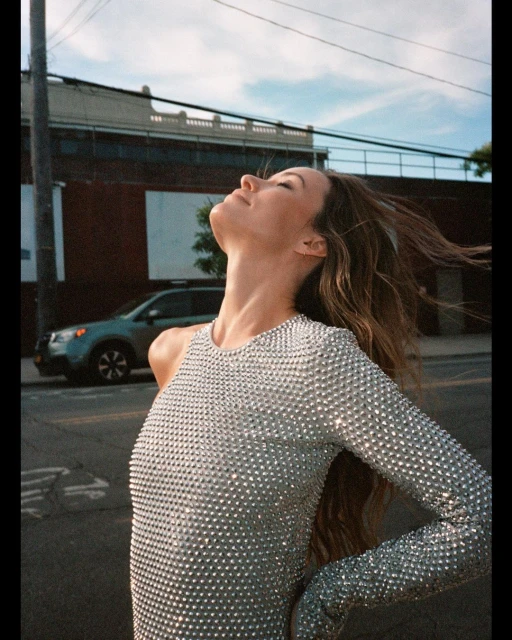 Adult female wearing a silver sequin dress poses outdoors with windblown hair, set against a building and a parked car under a blue sky.
