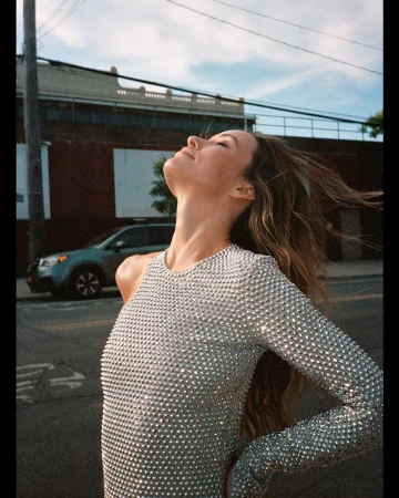 Adult female wearing a silver sequin dress poses outdoors with windblown hair, set against a building and a parked car under a blue sky.