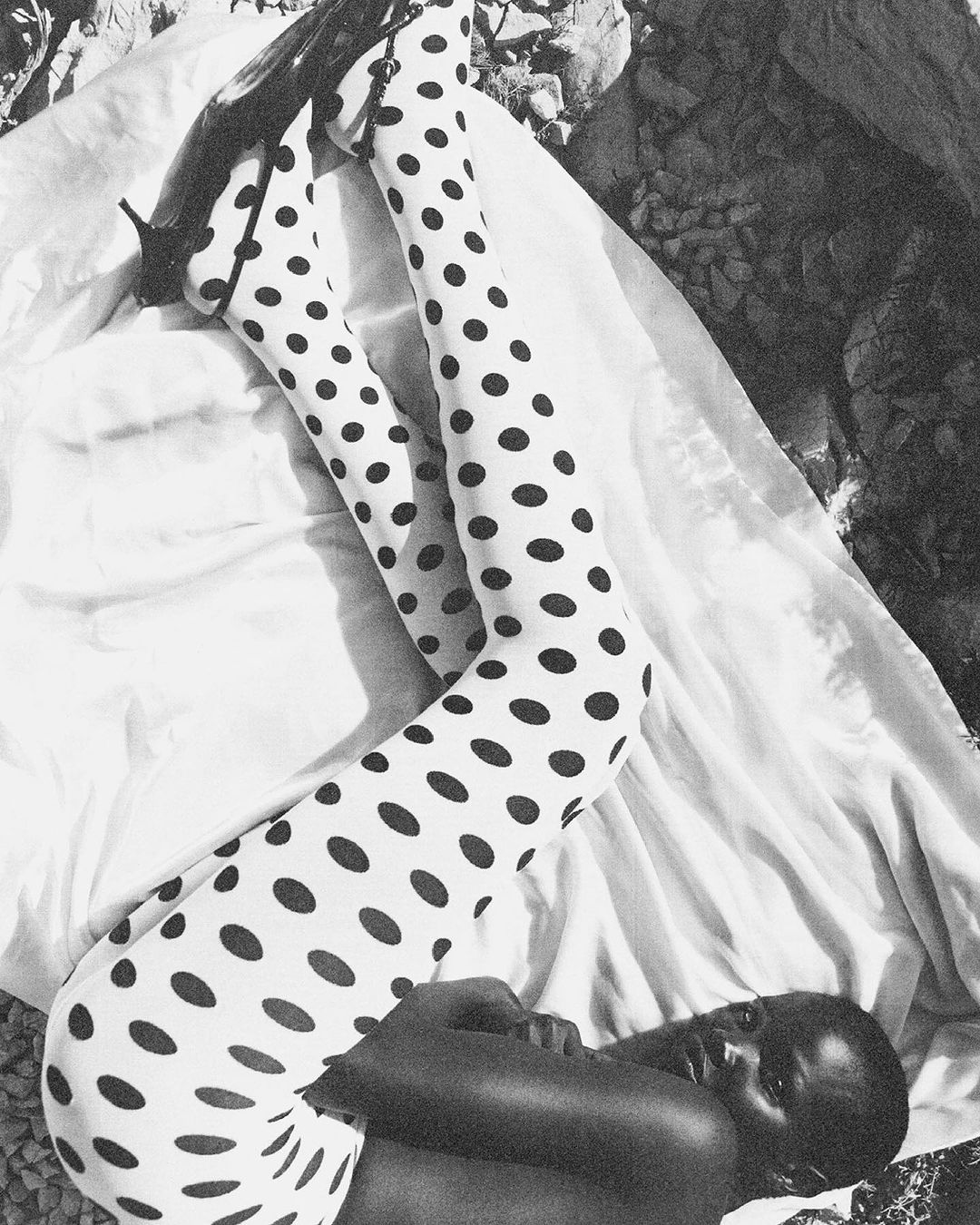 An adult woman poses on a white cloth outdoors wearing polka dot tights and black high heel shoes in a monochrome photograph.