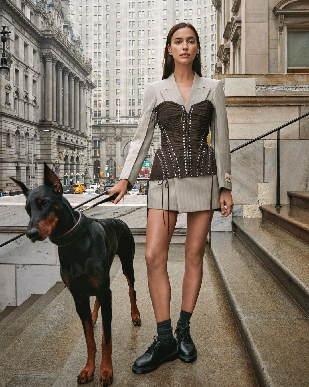 Fashionable woman with a Doberman dog in a city setting wearing a corset top, skirt, blazer, and black leather shoes on a city street.