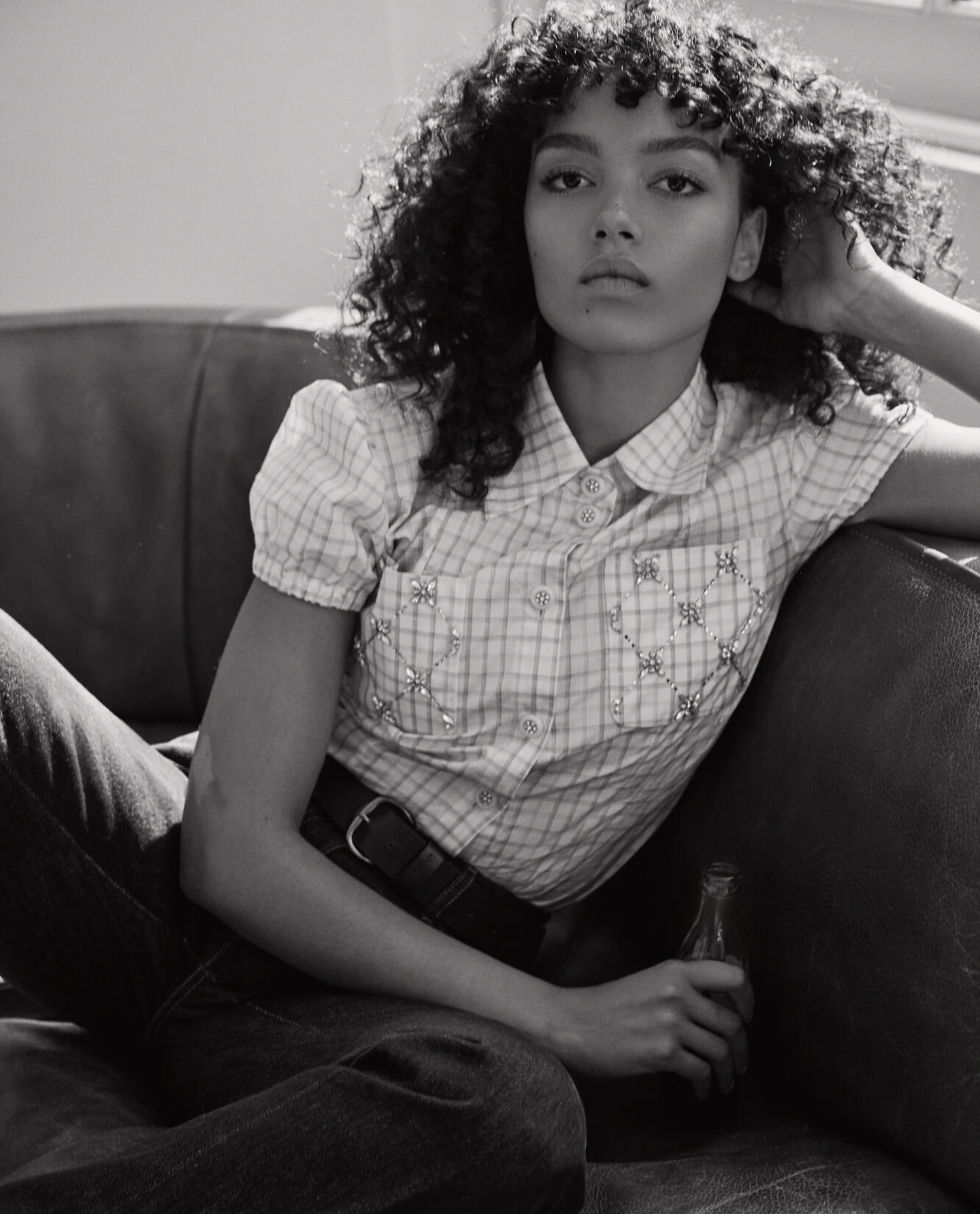 Black and white shot of a woman with curly hair sitting on a couch, wearing a plaid shirt, holding a vintage soda bottle.