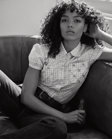 Black and white shot of a woman with curly hair sitting on a couch, wearing a plaid shirt, holding a vintage soda bottle.