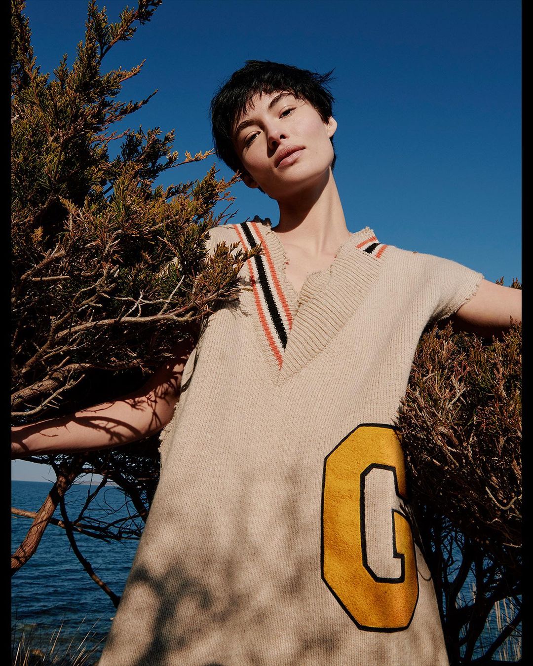 Woman poses in a beige sweater dress with a yellow letter patch, next to a green juniper bush with a bright blue sky and ocean backdrop.