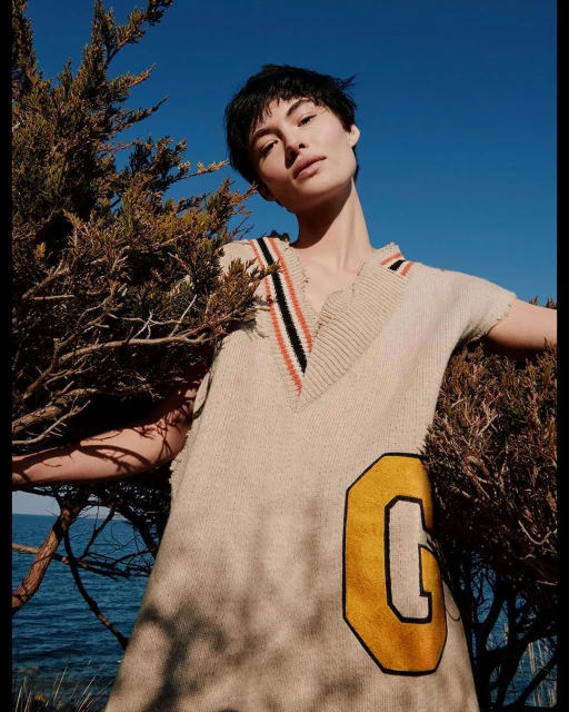 Woman poses in a beige sweater dress with a yellow letter patch, next to a green juniper bush with a bright blue sky and ocean backdrop.