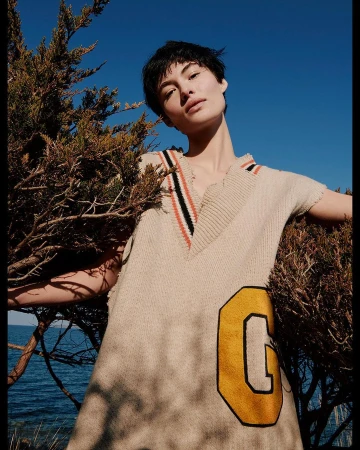 Woman poses in a beige sweater dress with a yellow letter patch, next to a green juniper bush with a bright blue sky and ocean backdrop.