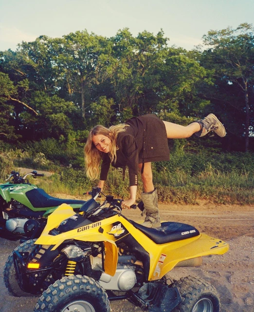A woman balances on a yellow ATV in an outdoor setting, with a green ATV visible in the background amid trees on a dirt road.