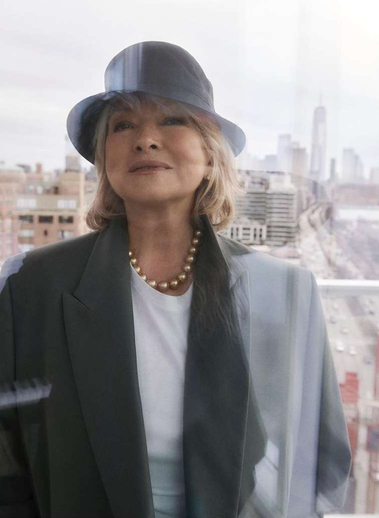 Woman in hat and blazer poses in front of city buildings. She wears a white top and gold jewelry, with a modern cityscape background.